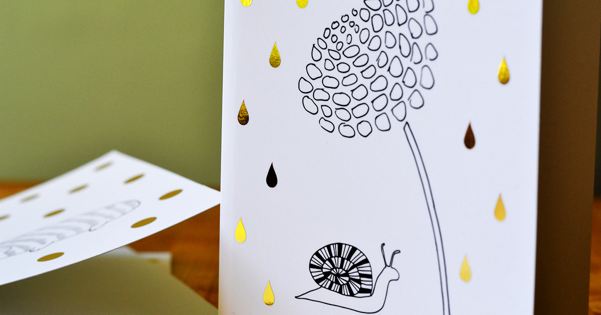 This is an image of a white card with a black and white image of a snail taking cover under a many petaled flower, while gold raindrops fall around it. The background of the photo is an olive green wall.